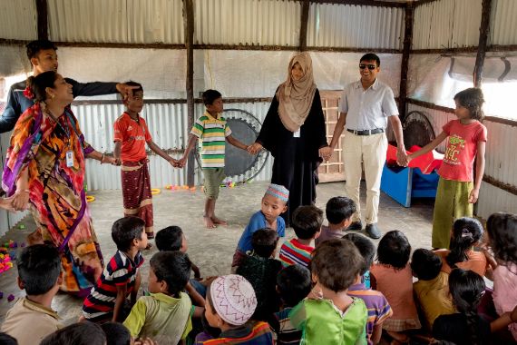 Kinder und Erwachsene spielen ein Kreisspiel und halten sich dabei an den Händen, andere Kinder sehen zu. Nach ihrer Vertreibung aus Myanmar leben sie in einem Flüchtlingscamp in Bangladesch.